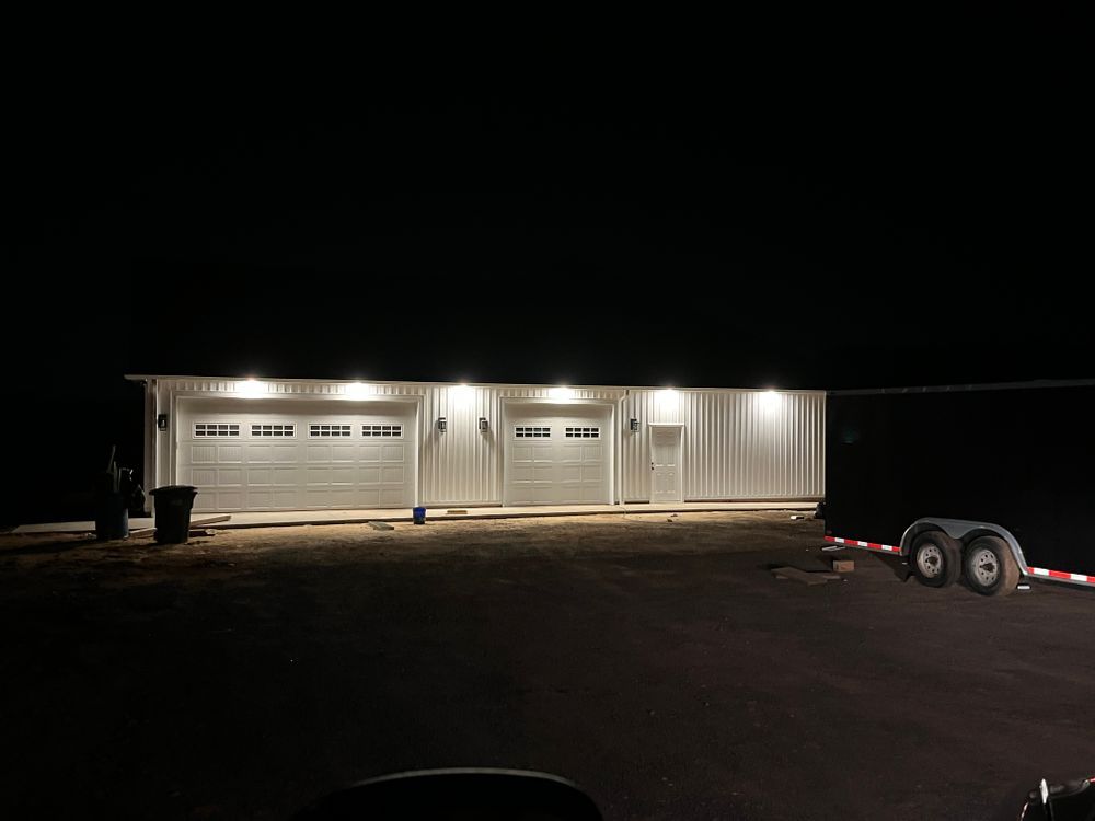 A white workshop with two garage doors and a side door, illuminated by outdoor lights at night on an unpaved lot.