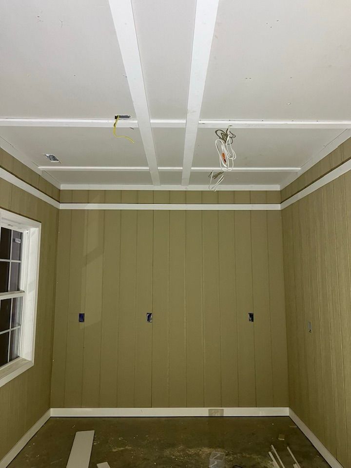A room undergoing renovation with vertical paneled, olive-green walls, a white coffered ceiling, and exposed wiring.
