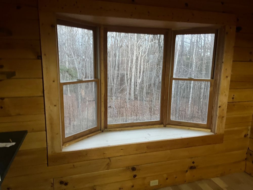 A large wooden bay window in a rustic cabin, showing a forest view through the glass.