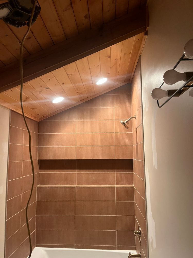 A bathroom shower with brown tiled walls and a slanted, wood-plank ceiling featuring two recessed lights.