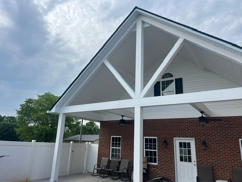 A large white triangular patio cover attached to a brick house with a concrete patio, white fence, and ceiling fans.
