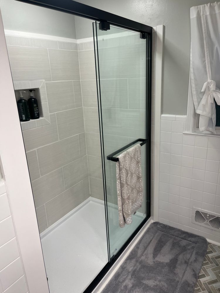 A walk-in shower with light gray tiled walls, a black-framed glass door, and a white floor, with a towel hanging on it.