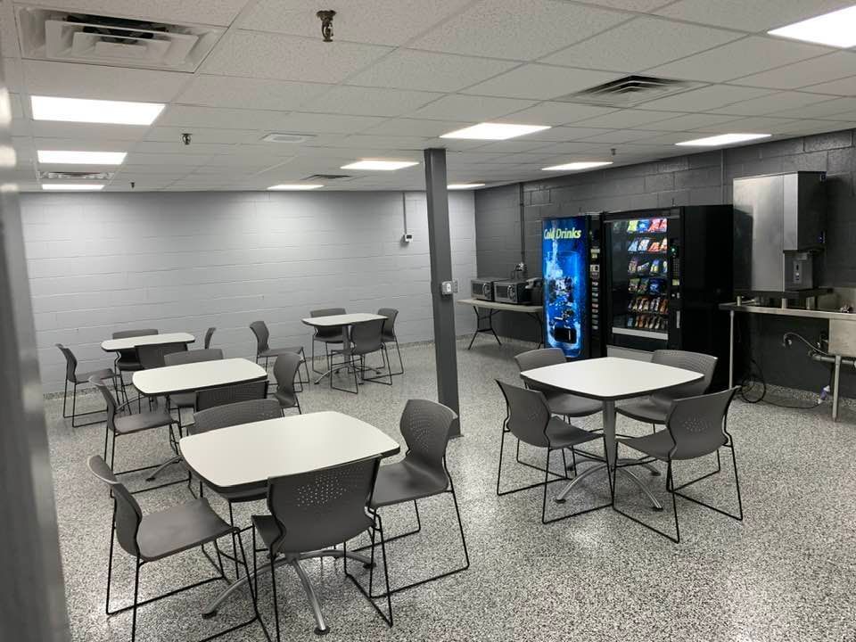 A breakroom with gray walls and speckled floors, featuring multiple square tables, gray chairs, & a bank of vending machines.
