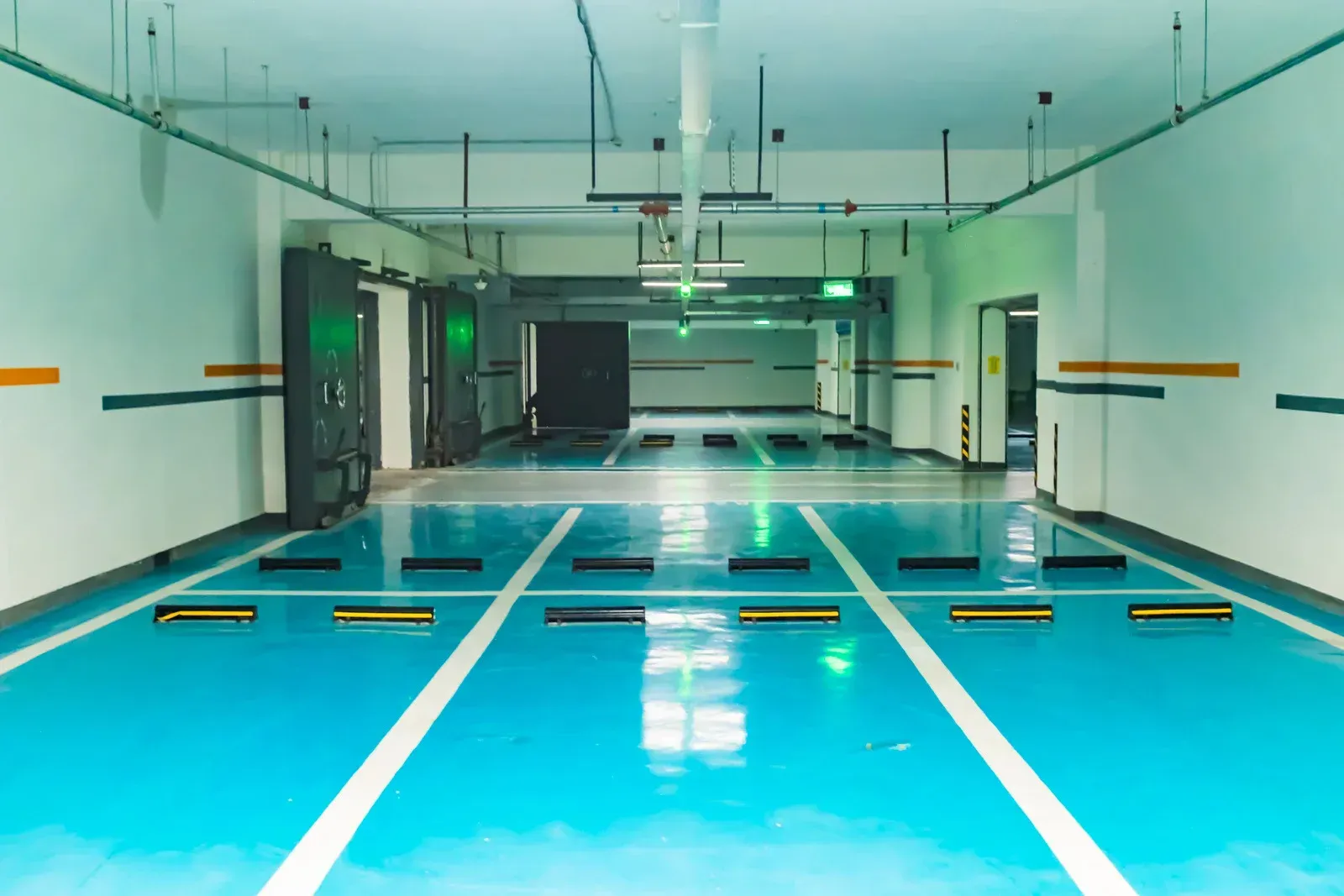 A brightly lit indoor parking garage with a glossy teal floor featuring white parking space markings and black wheel stops.