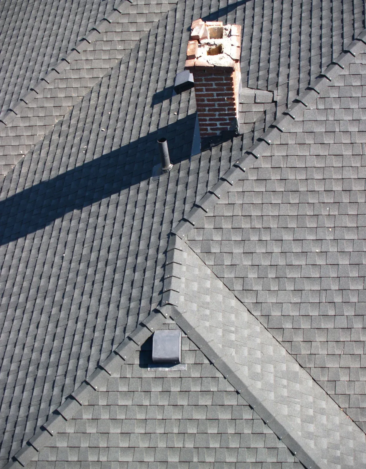 A high-angle view of a grey shingled roof featuring a brick chimney, a vent pipe, and a square roof vent.