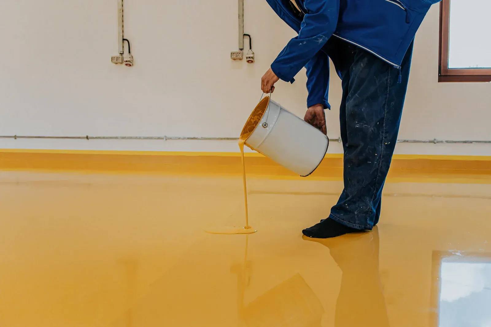 A person in blue workwear pours bright yellow epoxy resin onto a floor from a white bucket.