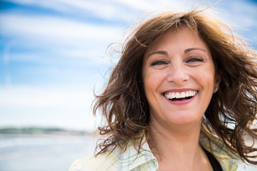 Woman with brown hair smiles broadly outdoors with a beach and blue sky in the background.