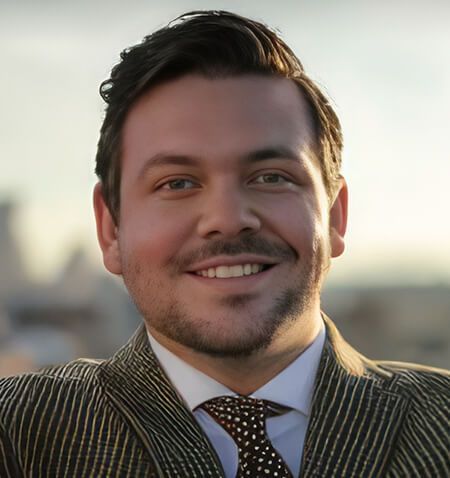 Man smiling, wearing a striped suit and polka-dot tie, with city backdrop.