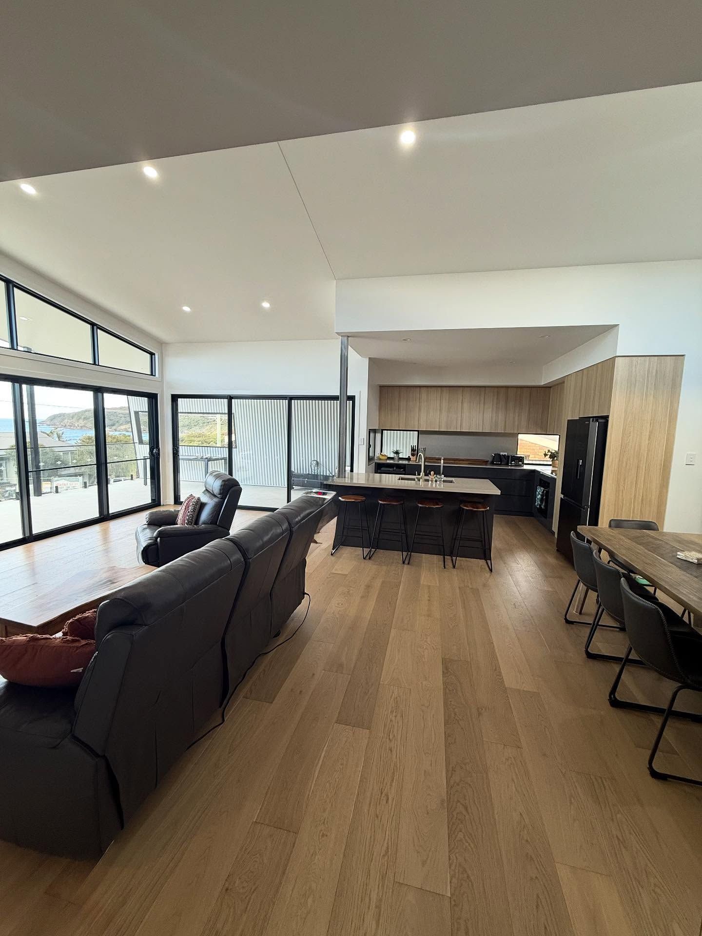 Open-plan living space with light wood floors, a kitchen island, and a dark leather sofa. — Barrington Coast Homes in Tallwoods, NSW