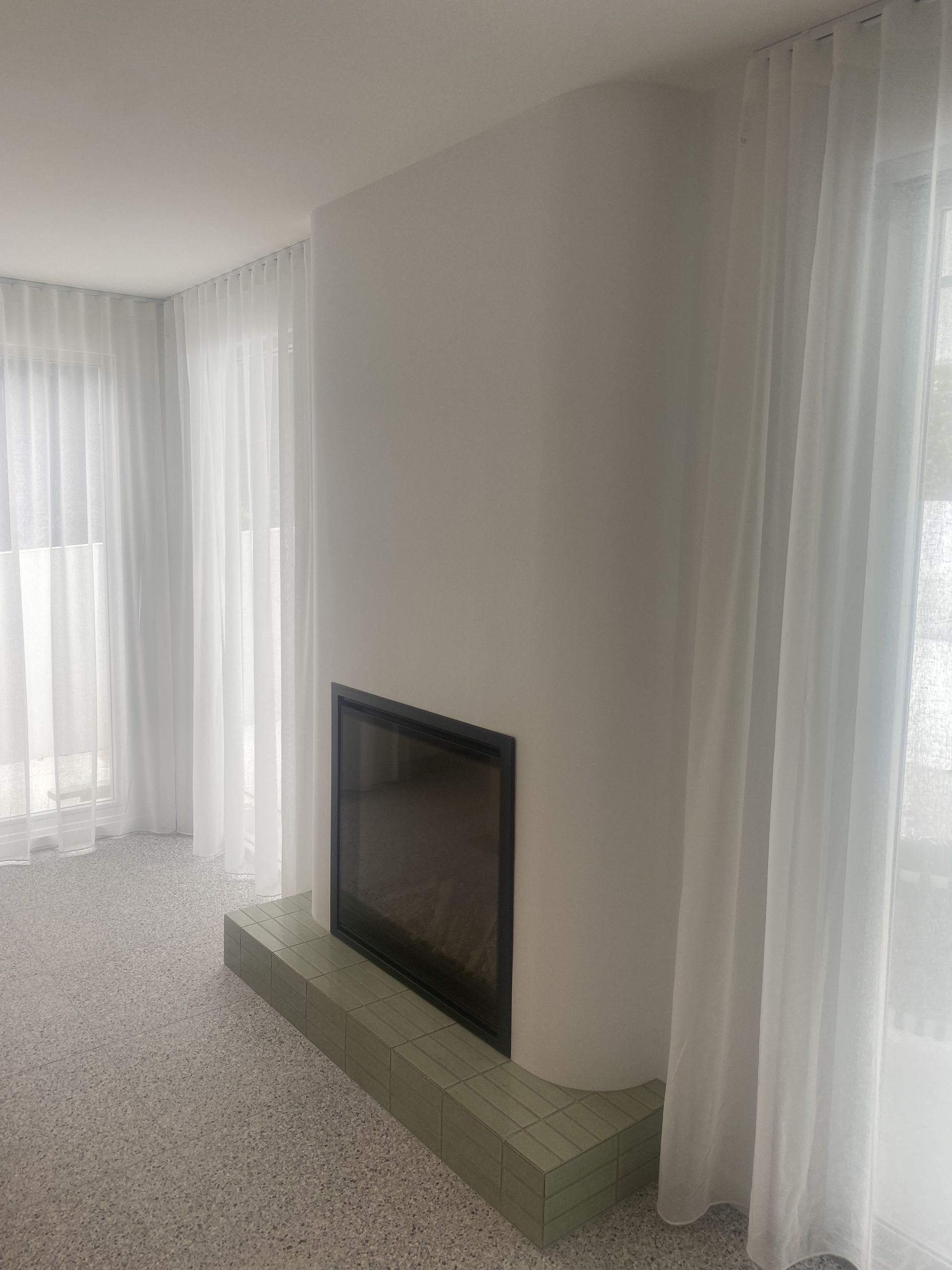 Fireplace with dark glass and light green base, flanked by sheer white curtains and bright terrazzo flooring.