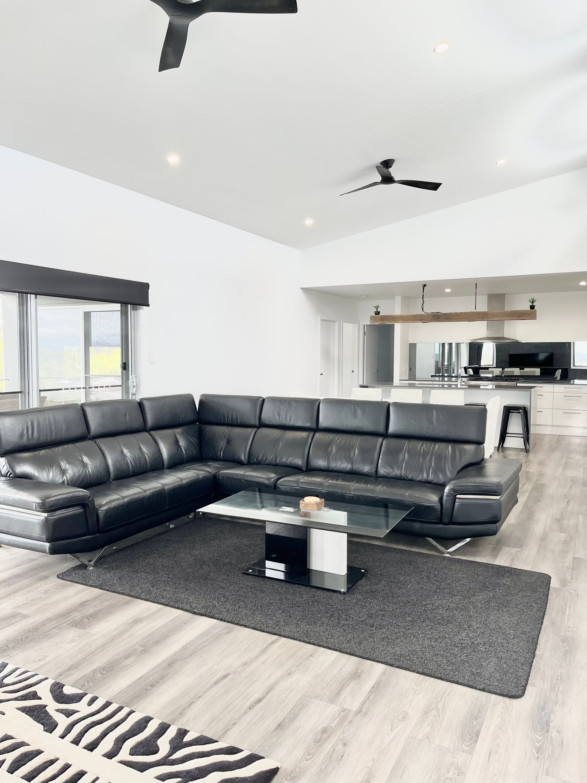 Modern living room with black sectional sofa, rug, and glass coffee table. White walls, wood floor, open kitchen — Barrington Coast Homes in Tallwoods, NSW