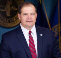 Man in a navy suit and red striped tie, smiling, in front of a county seal and flags.