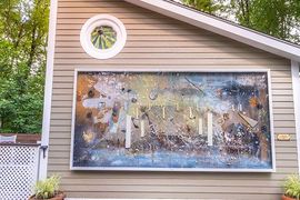 A large, framed abstract mural with blue and earthy tones hangs on beige siding beneath a circular window.