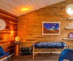 Cozy wood-paneled room with a bench, glowing lamp, framed artwork, and a blue chair