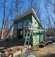 Small green cabin with ladders on a wooded site under a bright blue sky