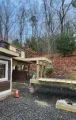 Outdoor house deck and gravel yard beside a wooded hillside, with a covered patio and construction materials.