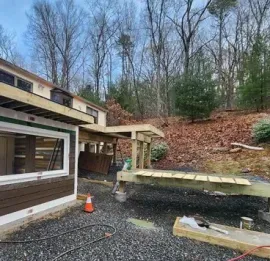 Construction of a wooden deck frame attached to a house with brown siding, situated on a gravel lot near a wooded area.