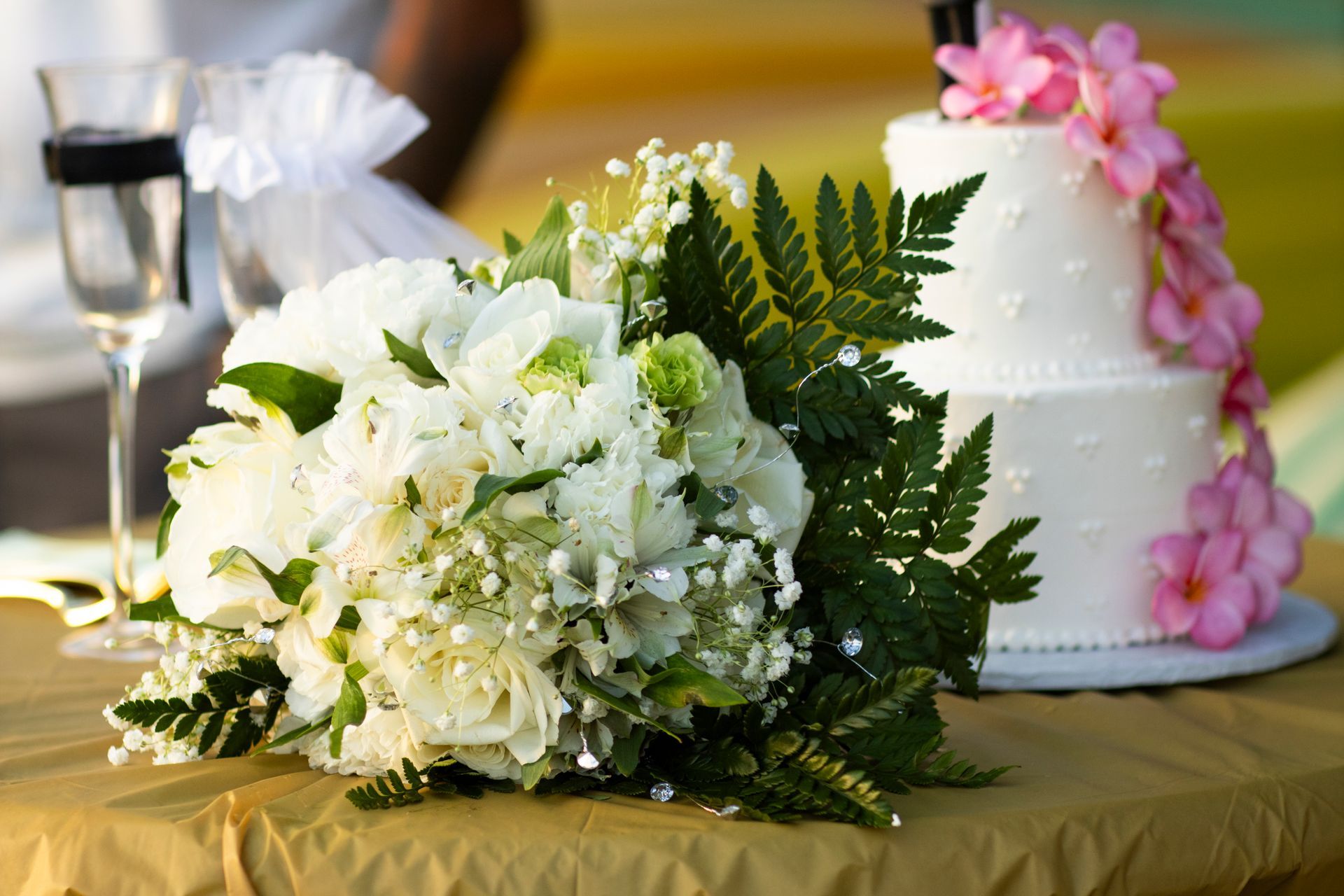 Wedding bouquet with white flowers and ferns on a gold tablecloth next to a cake decorated with pink flowers.