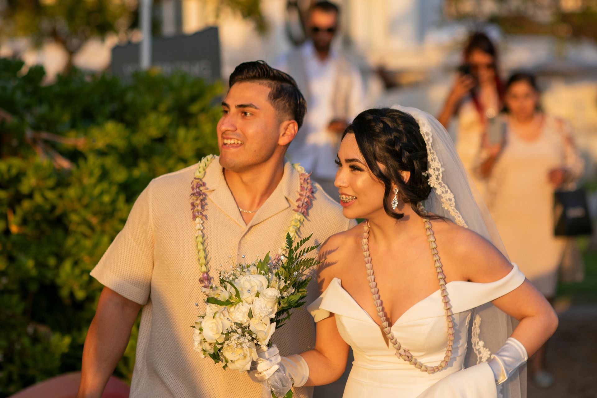 Bride and groom smiling, walking outside during golden hour; bride in white dress, groom in beige shirt.