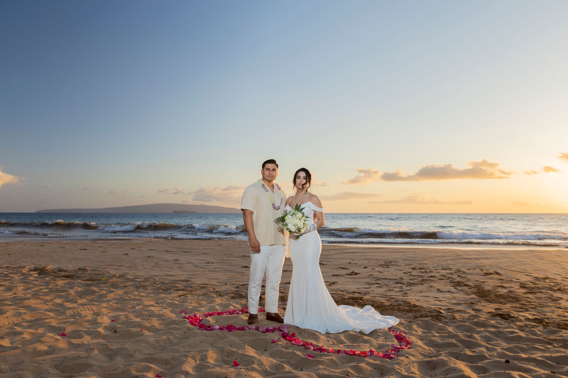 Couple stands on a beach, framed by a rose petal heart at sunset.
