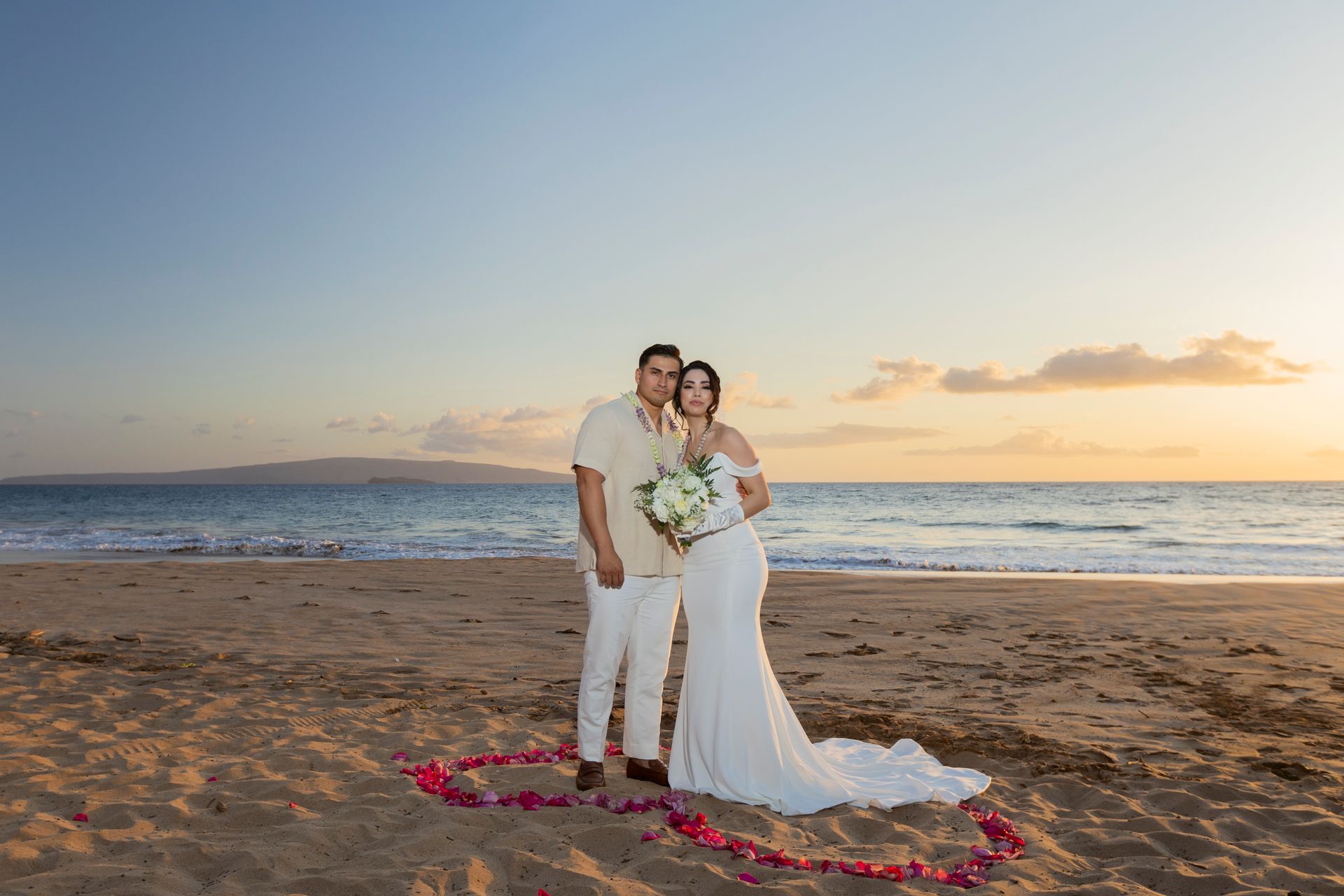Newlyweds stand on a beach at sunset, framed by a heart of flower petals.