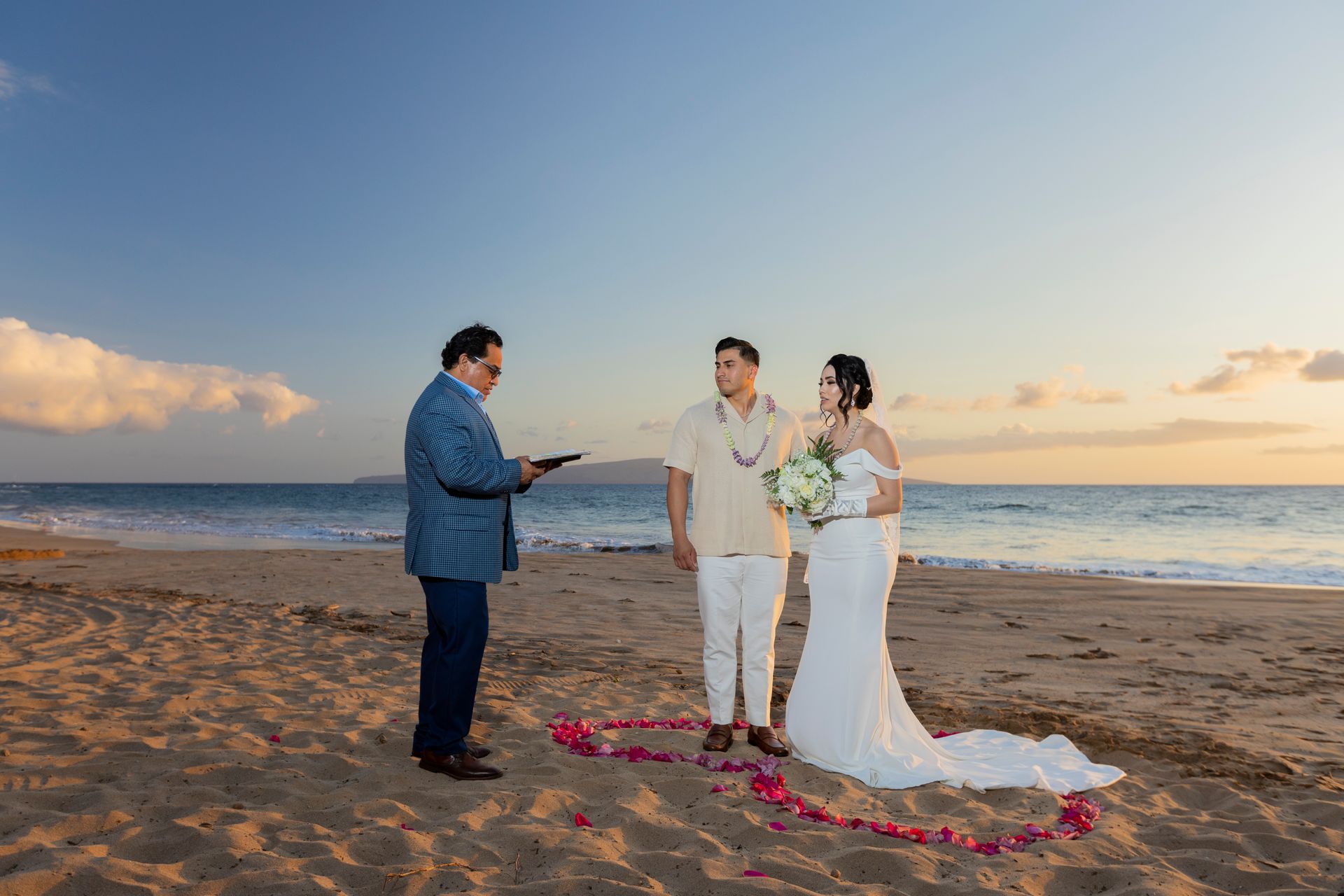 Wedding ceremony on a beach: couple with officiant, floral heart on sand, sunset.