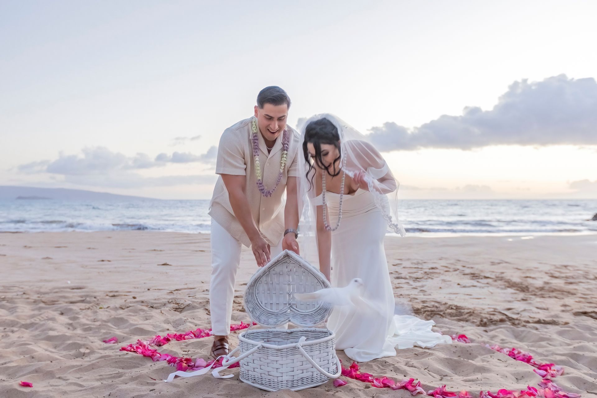 Couple at beach wedding: man and woman pouring sand into a decorative chest. Pink rose petals surround them.