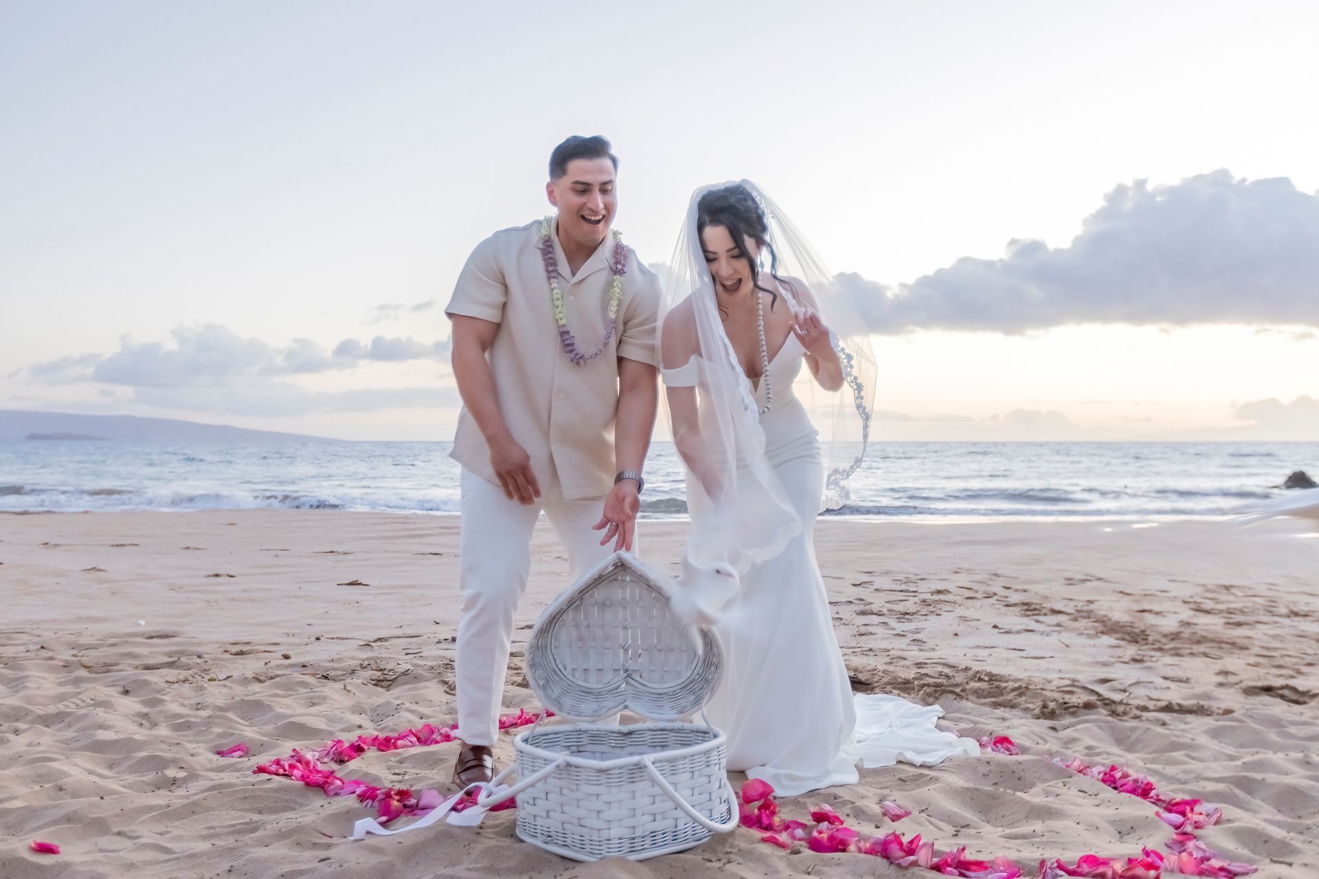 Wedding couple opens heart-shaped basket on beach. Man points, woman looks surprised. Pink rose petals surround. Ocean and sky in background.