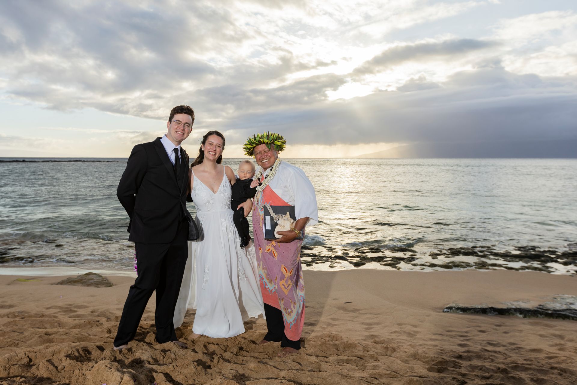Wedding couple with officiant on a beach at sunset; man in suit, woman in white dress, officiant in traditional wear.