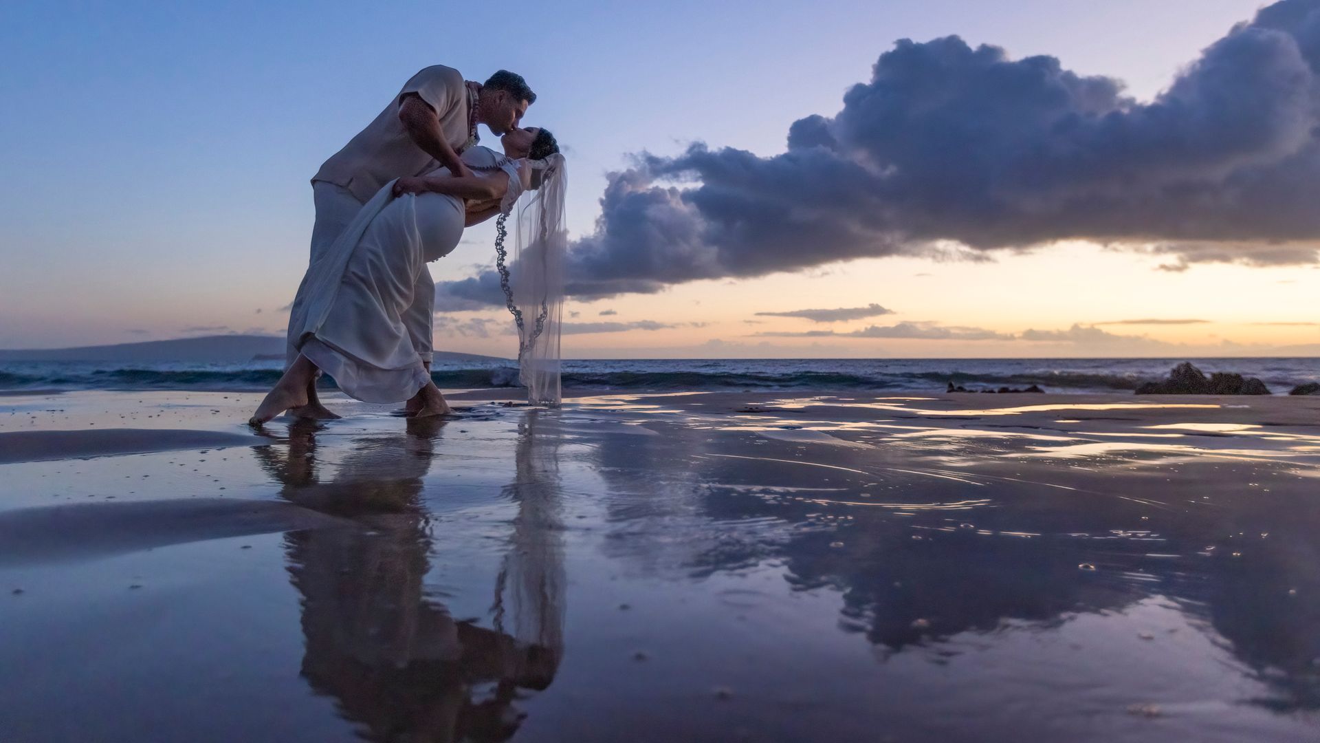 Couple kissing on a beach at sunset, reflection in wet sand.