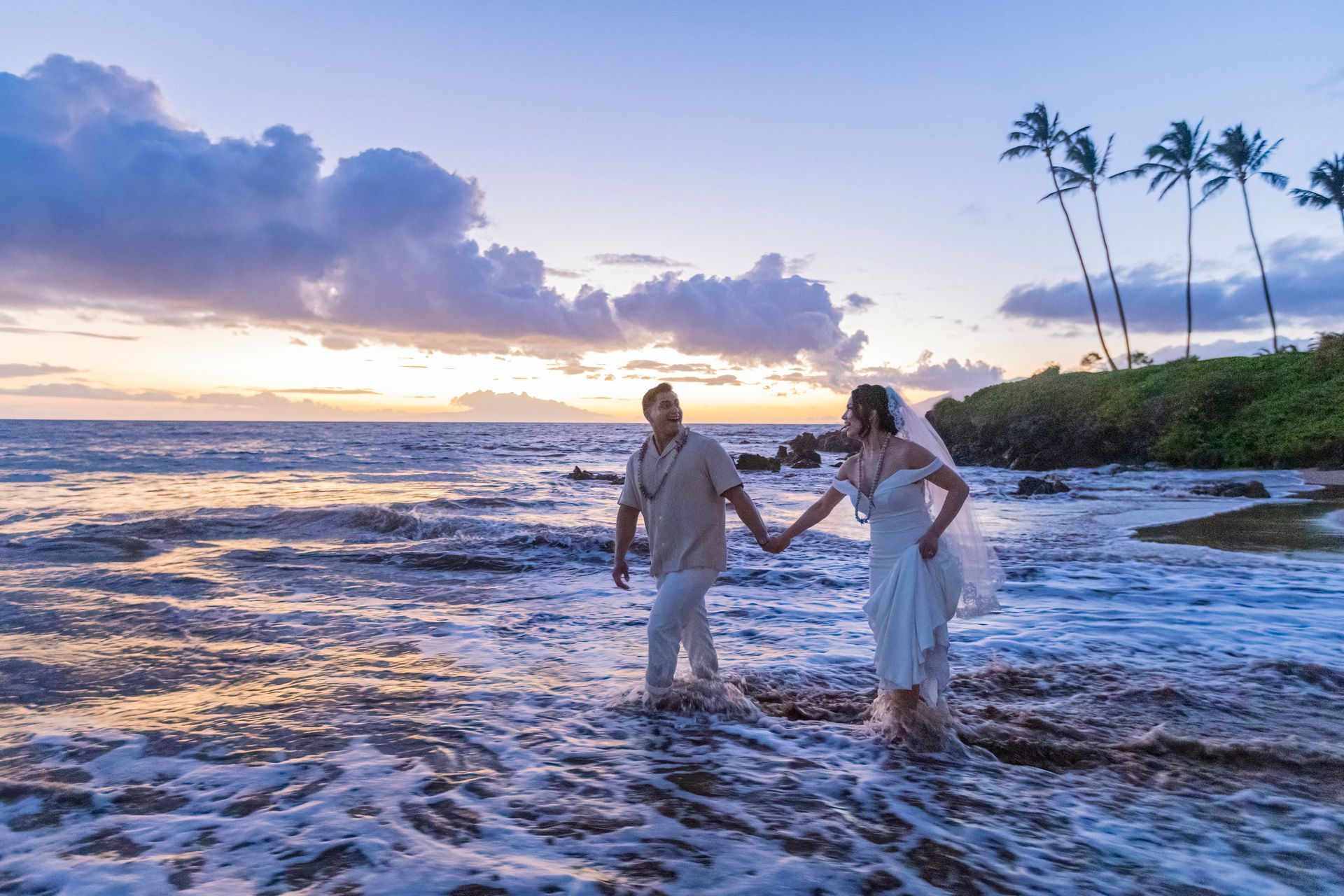 Bride and groom walk hand-in-hand in ocean waves at sunset, with palm trees in the background.