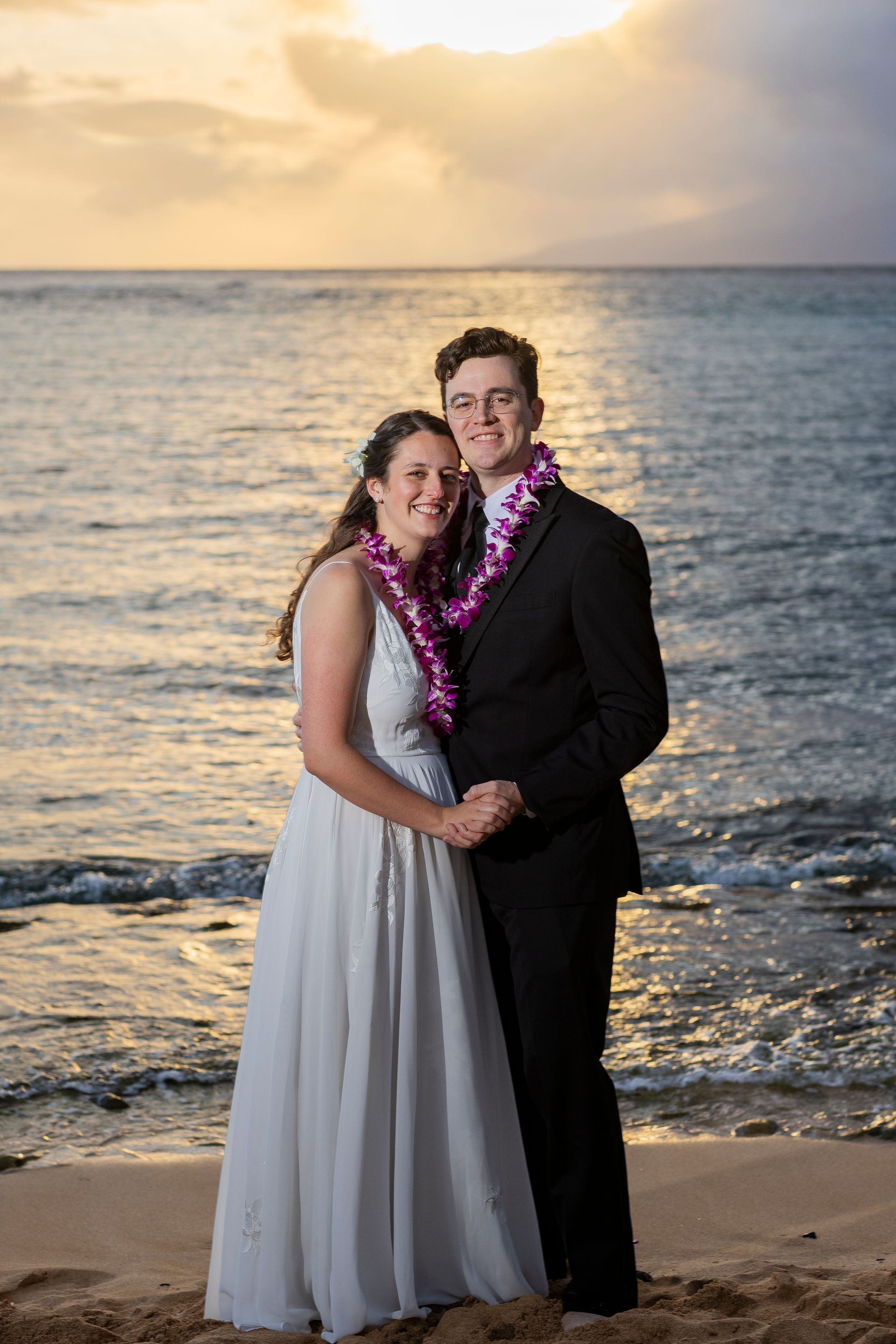 Newlyweds on a beach at sunset, holding hands, wearing leis, she in a white dress, he in a suit.