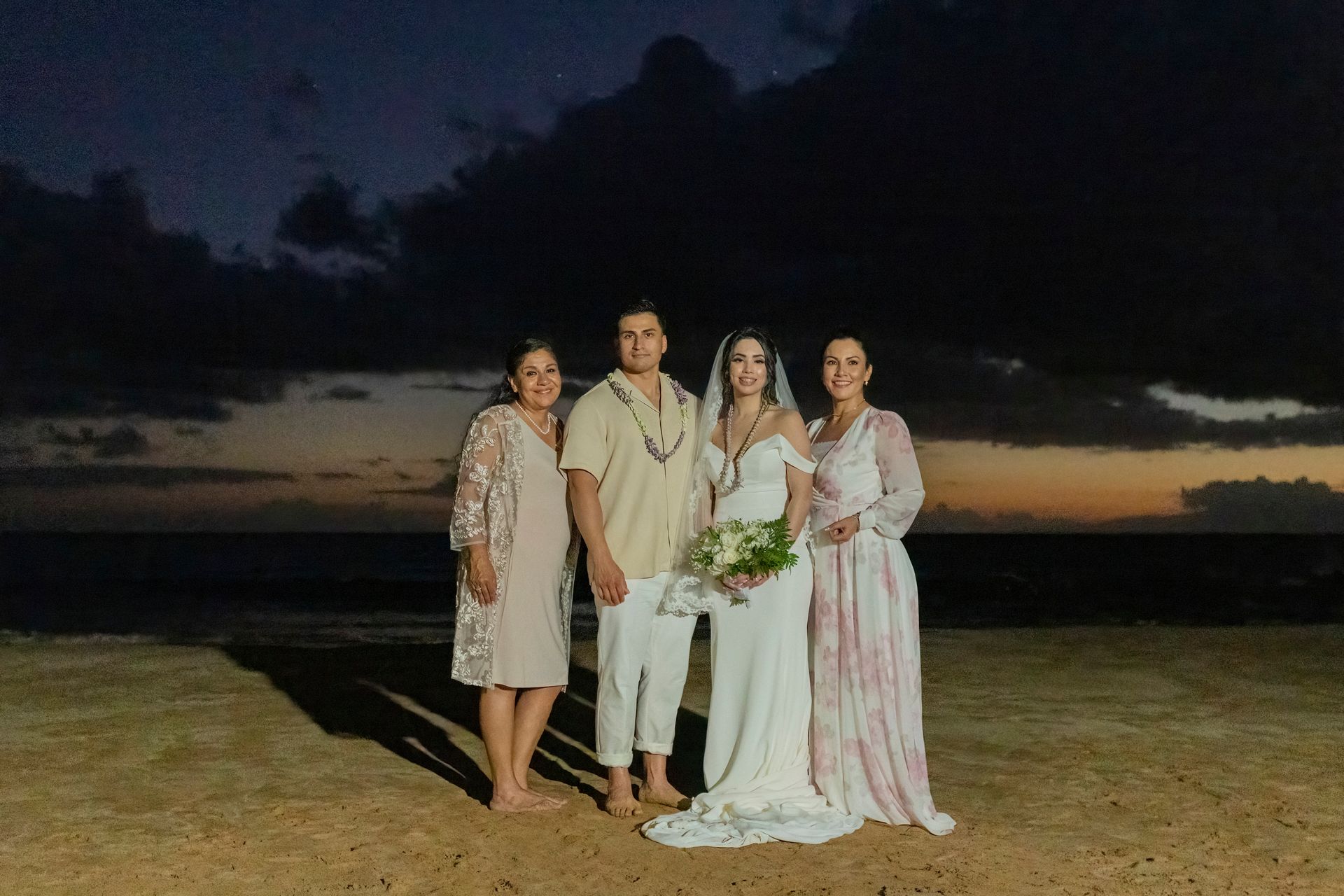 Bride and groom with two women on a beach at sunset. The bride holds a bouquet and wears a white dress.