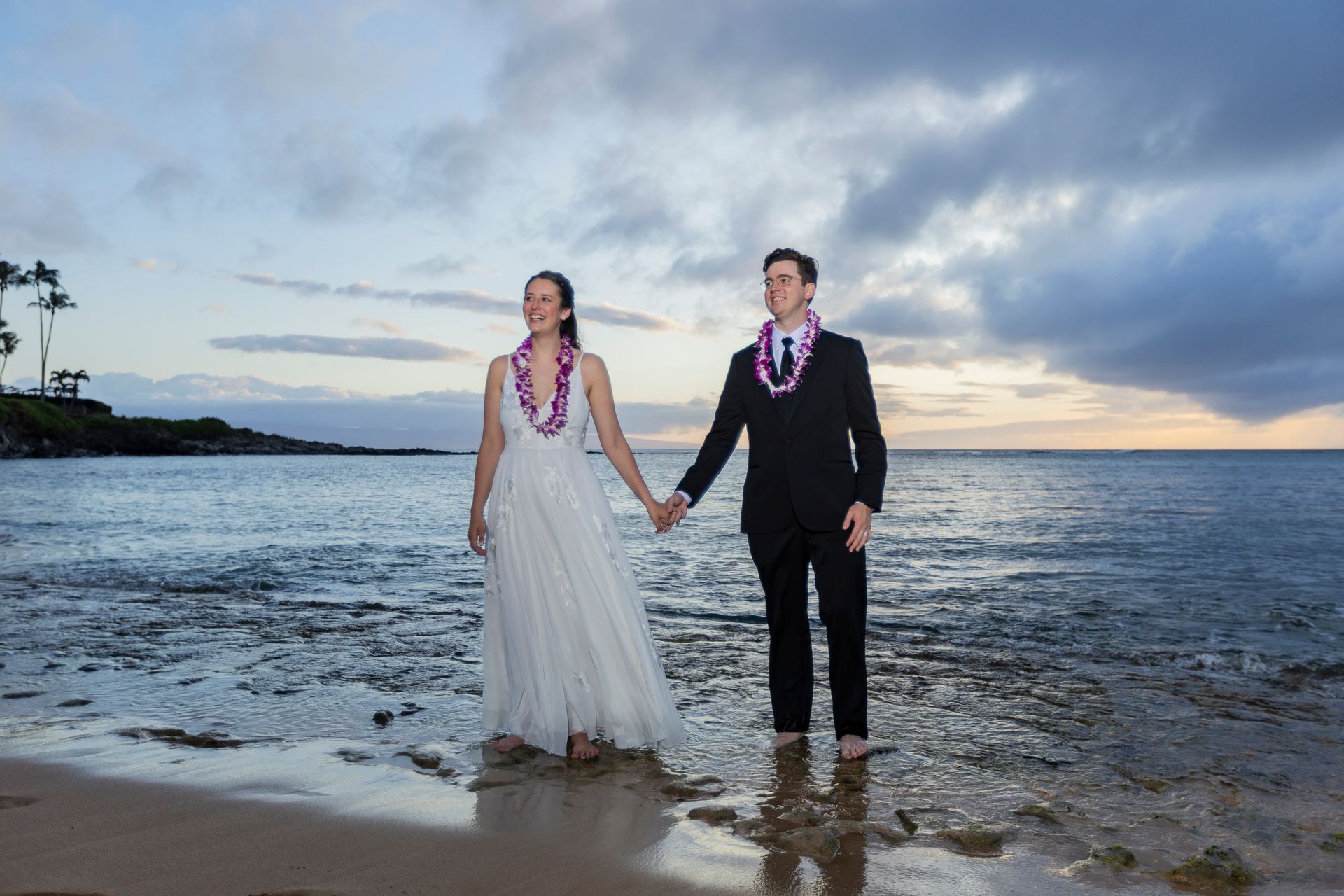 Bride and groom holding hands on a beach, wearing leis. Ocean and cloudy sky in the background.