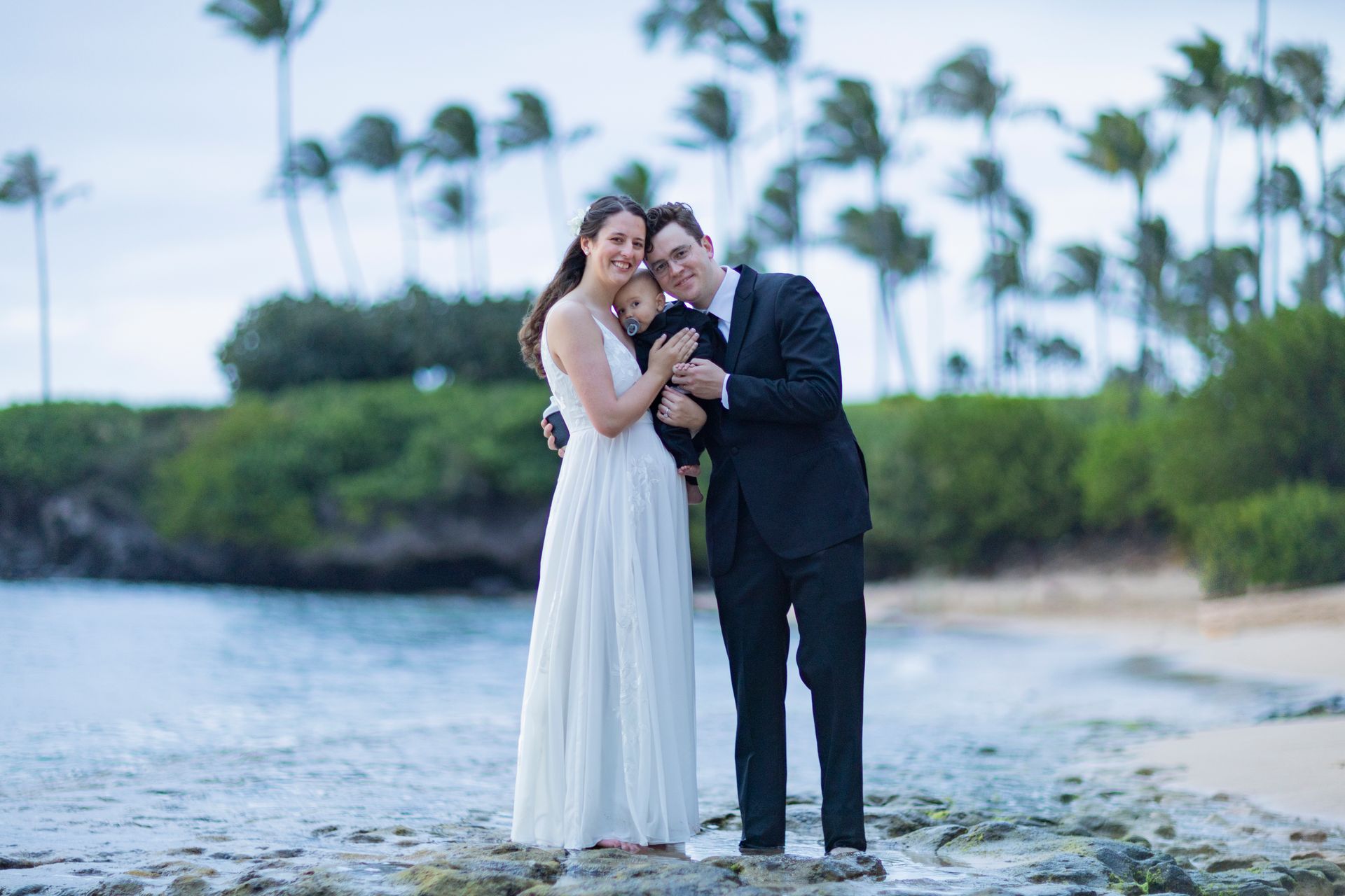 Newlyweds and baby pose on a beach; woman in white dress, man in suit, holding the baby.