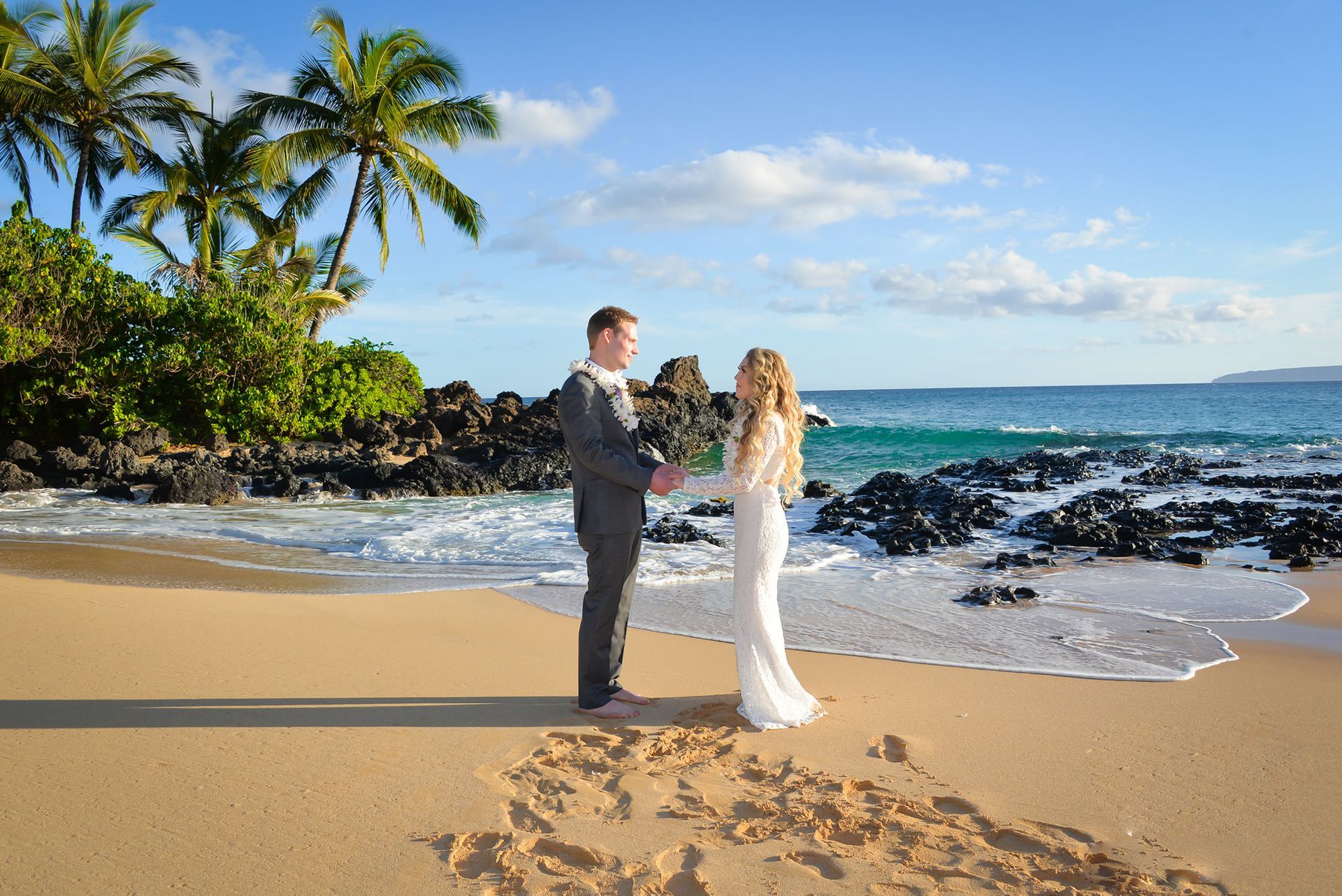 Couple holding hands on a sandy beach, with palm trees and ocean in the background.
