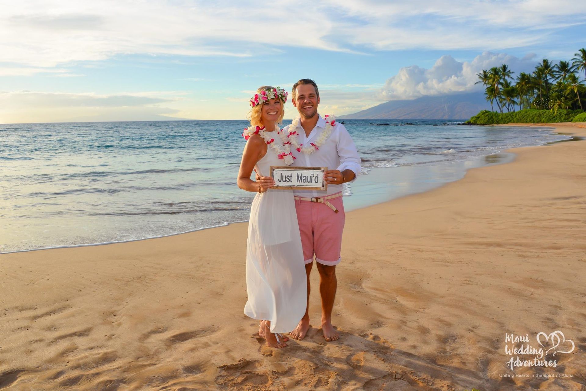 Couple on beach, bride in white dress, groom in pink shorts, holding a framed sign, sunset.