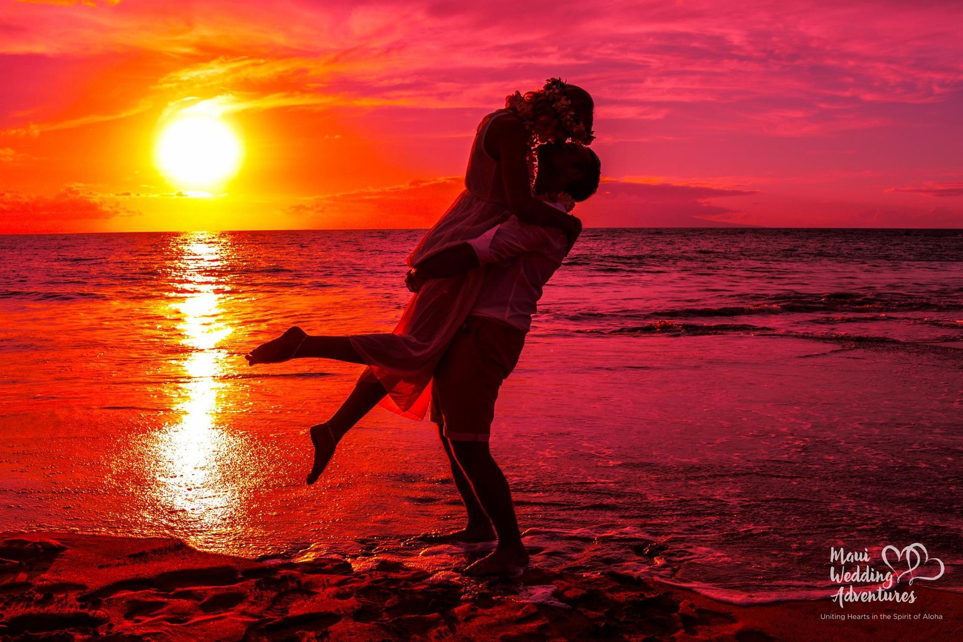 Couple embracing on a beach as a fiery sunset reflects on the water.