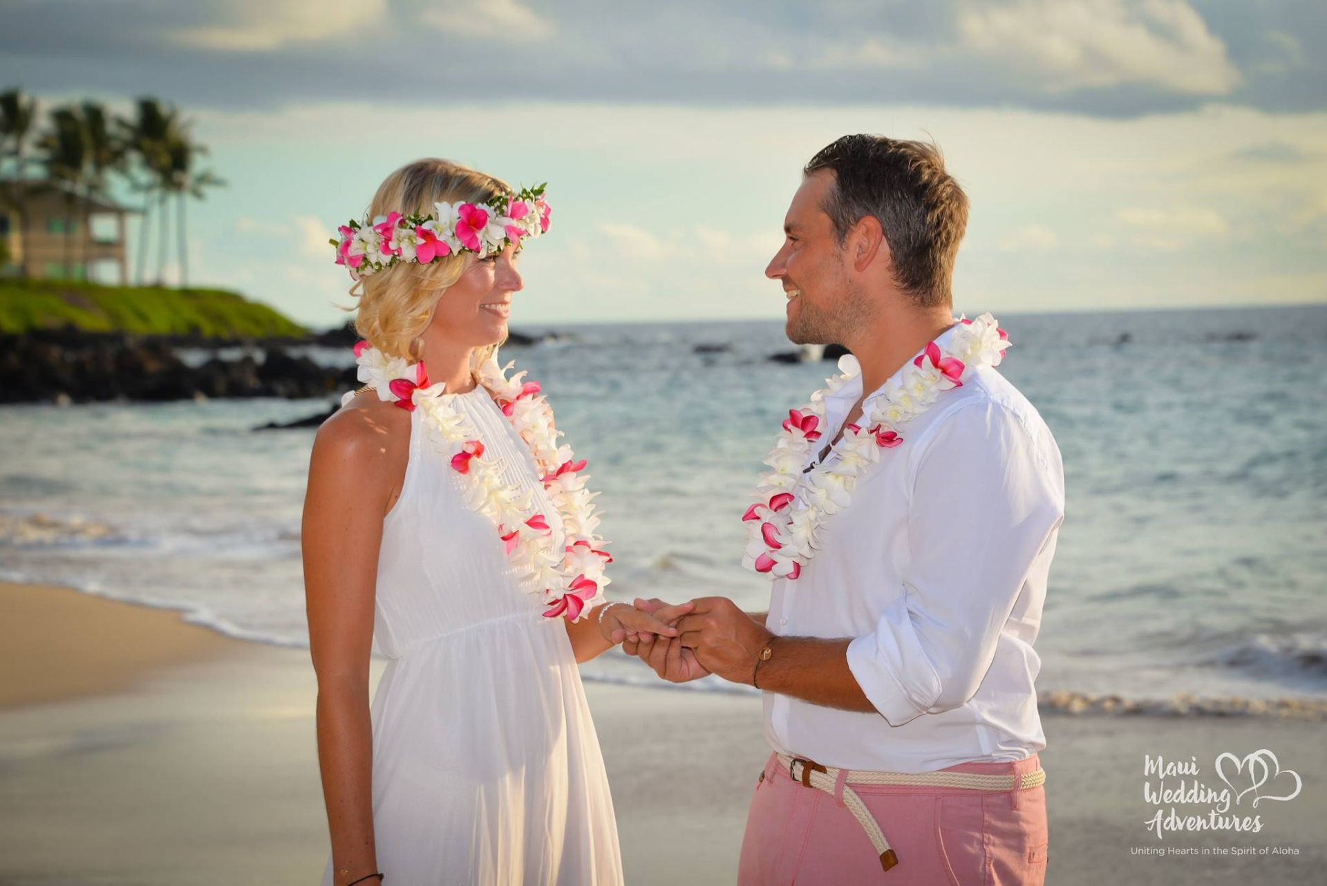 Couple on beach, bride in white dress and flower crown, groom in pink pants, holding hands. Ocean in background.