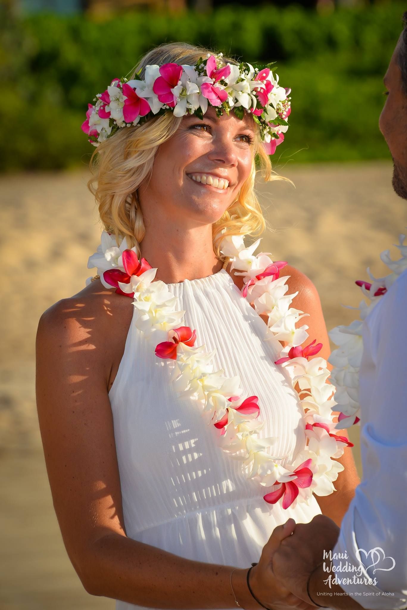 Bride in white dress and flower crown, smiling, with lei, beach setting.