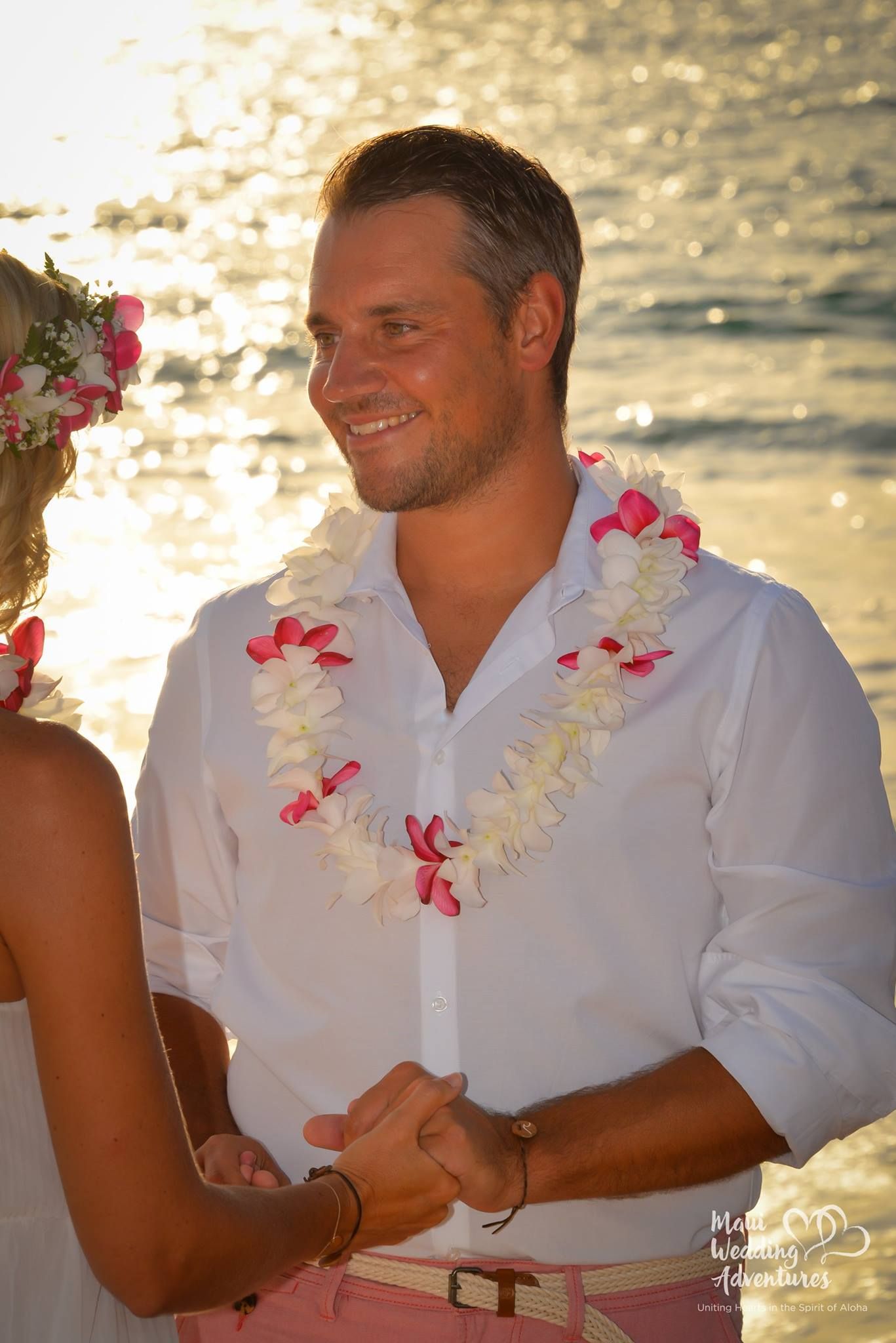 Man in white shirt, wearing a lei, smiling at a wedding on the beach, holding hands.