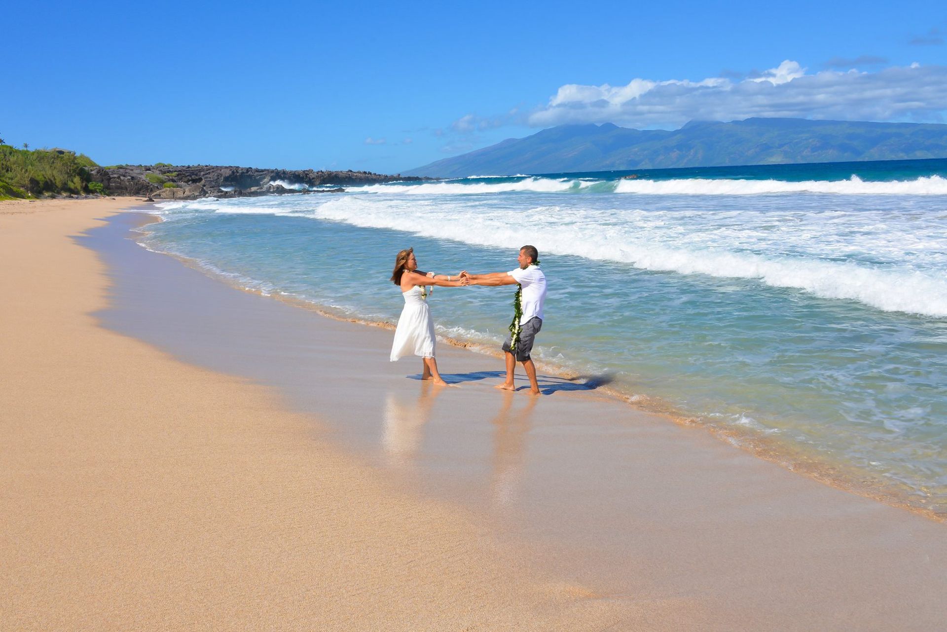 Couple holding hands, dancing in the shallow waves on a sandy beach under a bright blue sky.
