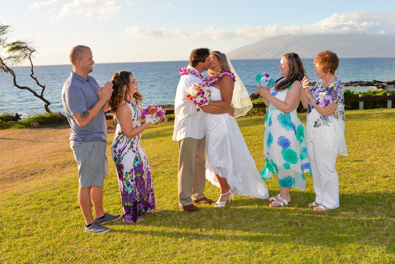 Wedding on a grassy hill. Bride and groom kissing surrounded by wedding party near ocean.