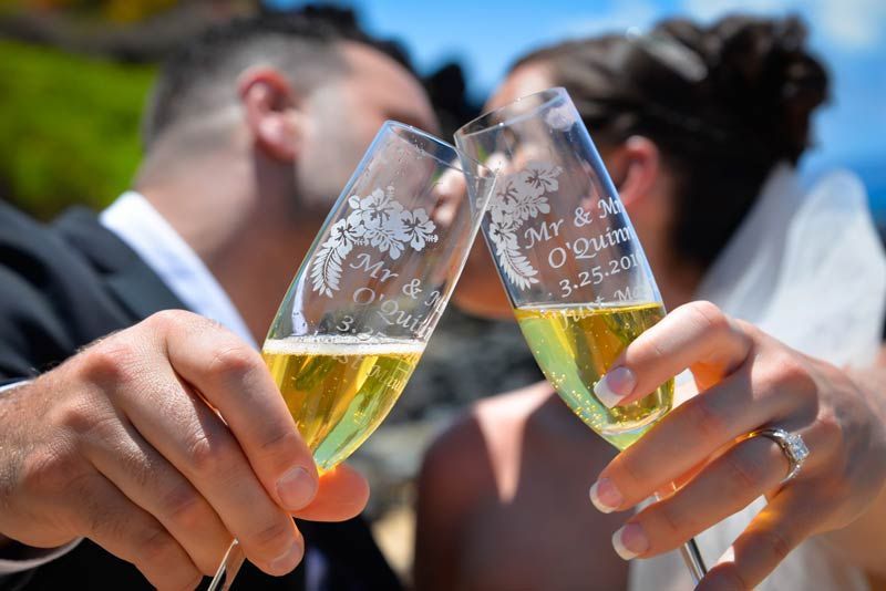 Newlyweds clinking champagne glasses, kissing. Clear glasses etched with names and date, outdoors with blue water.