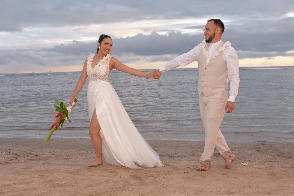 Bride and groom holding hands on beach, laughing. Cloudy sky and ocean in background.