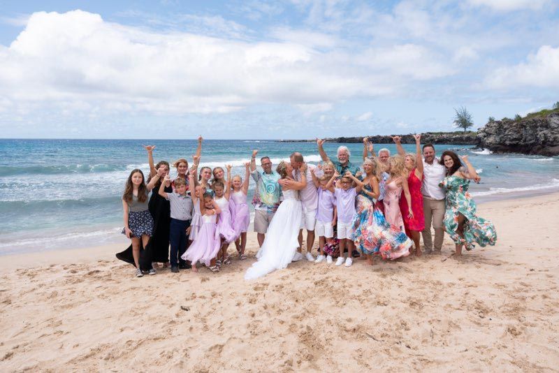 Wedding group on beach, bride & groom in center, arms raised. Ocean background, blue sky, sunny day.