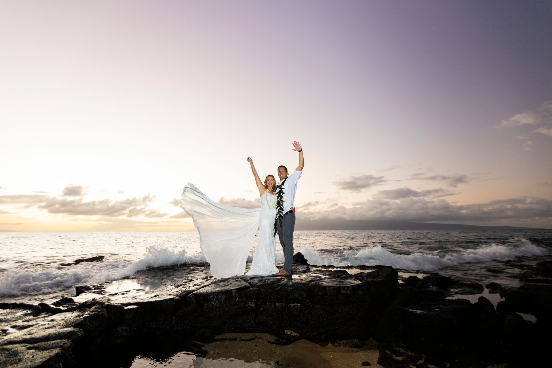 Newlyweds stand on rocks at sunset, arms raised in celebration. Waves crash around them.