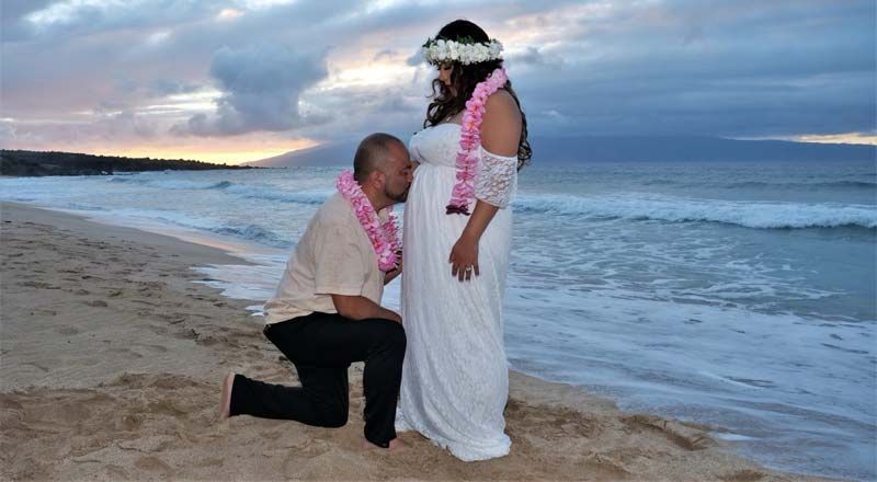 Man kneeling to kiss pregnant woman's belly on a beach; both wearing leis.