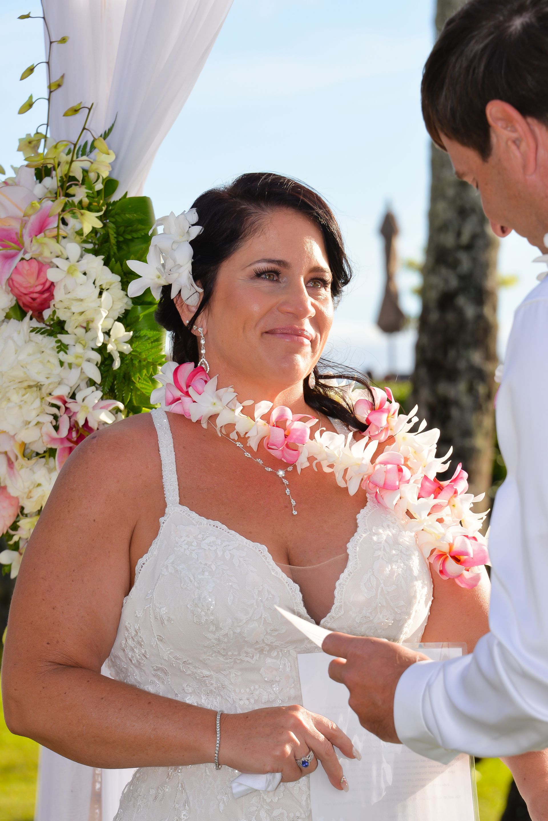 Bride in white dress, floral lei, looks at groom reading vows during outdoor wedding ceremony.