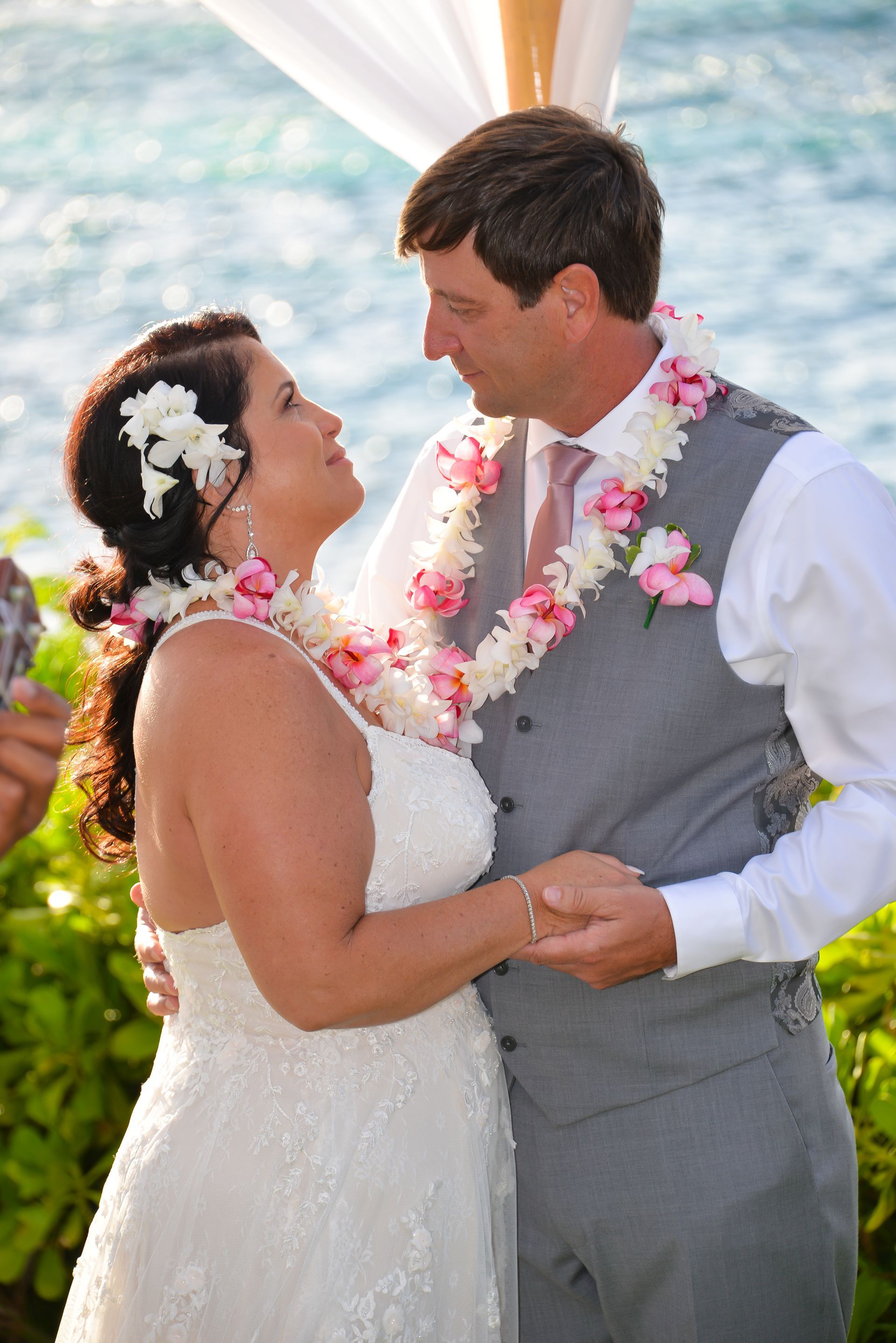 Bride and groom exchange vows outdoors by the ocean, both wearing flower leis.